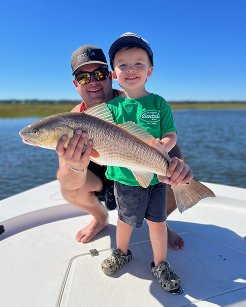 A smiling child in a green shirt and shorts proudly holds a freshly caught red drum fish with the help of an adult behind him on a boat. The adult, wearing sunglasses and a cap, steadies the fish while kneeling. The background shows calm coastal waters and clear blue sky on a sunny day. Provided photo/CRD.